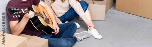 panoramic crop of woman sitting on floor with man playing acoustic guitar near boxes  relocation concept