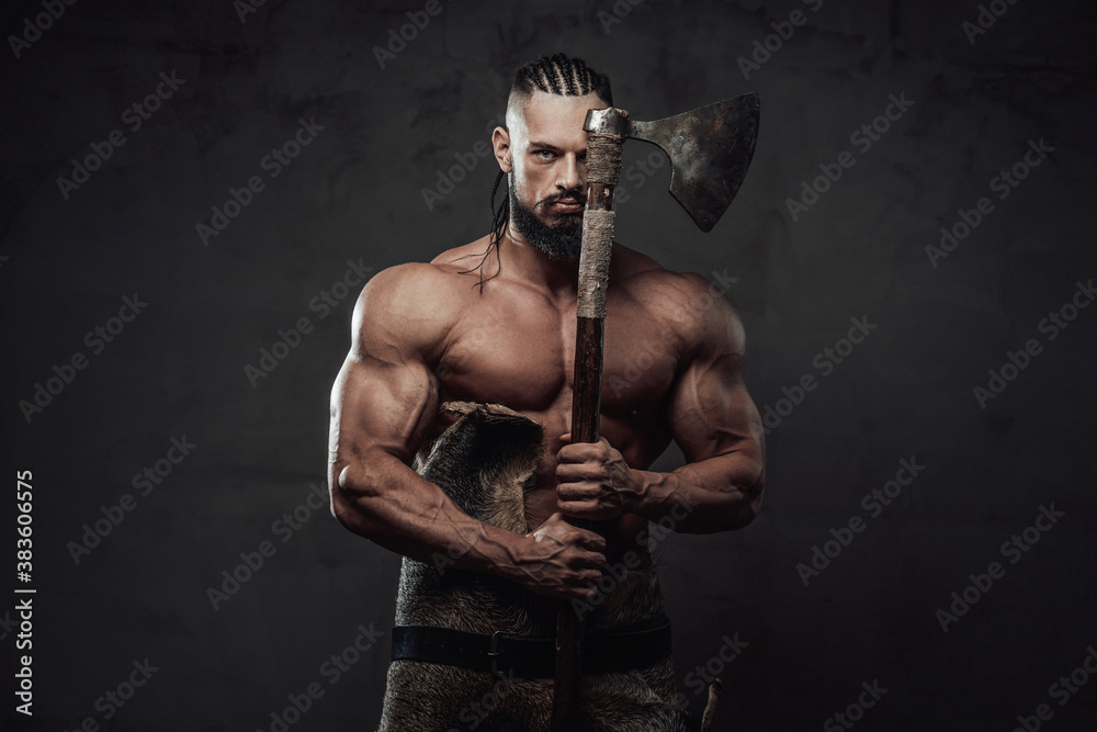 Brutal and powerful viking warrior with dreadlocks and beard posing with axe which covers half of his face in dark background.