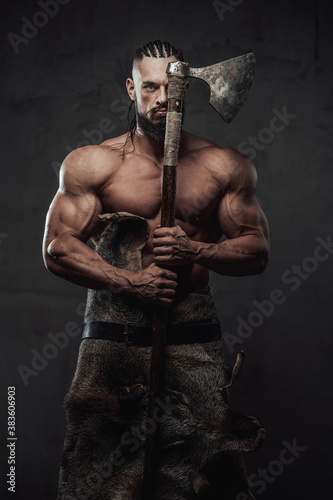Ancient muscular viking staying with naked torso and dreadlocks holding his axe hiding half of his face in dark background. © Fxquadro