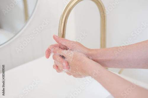 Washing hands with soap at a bathroom sink cleaning under running water under brushed brass faucet tap mixter basin with white tiles and mirror lit by natural light from nearby window foam soap santiz photo