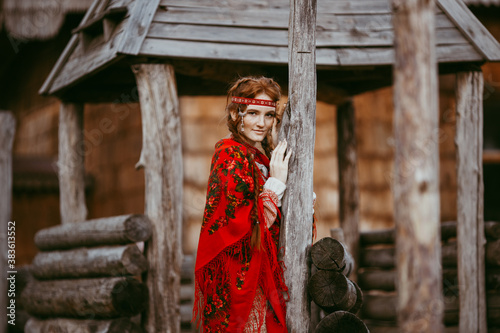 A beautiful Slavic girl with long blonde hair and brown eyes in a white and red embroidered suit is sitting next to a wooden house.Traditional clothing of the Ukrainian region. © Aleksei Zakharov