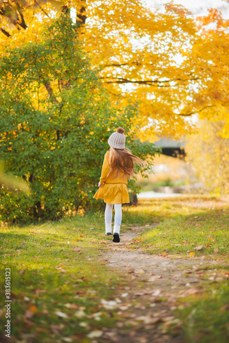 A little girl in a yellow dress and a brown sweater runs along the path in the autumn Park. A girl with red hair and a yellow dress runs along a path in an autumn Park. Autumn photo shoot of a girl wi