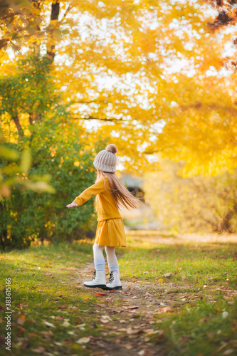A little girl in a yellow dress and a brown sweater is spinning on the path in the autumn Park. A girl with red hair and a yellow dress runs along a path in an autumn Park. Autumn photo shoot of a gir