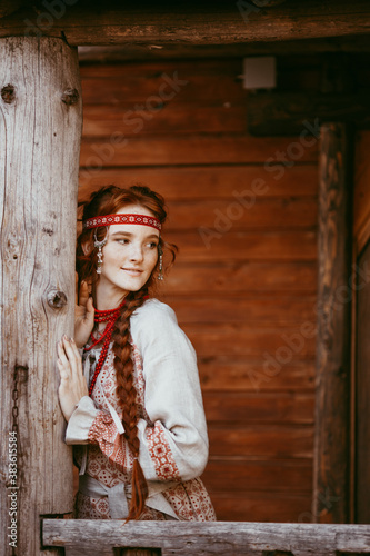 A beautiful Slavic girl with long blonde hair and brown eyes in a white and red embroidered suit stands on the porch of a house.Traditional clothing of the Ukrainian region. © Aleksei Zakharov