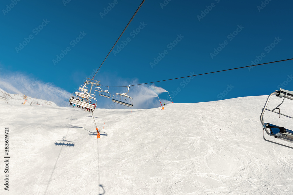 Scenic panoramic view of alpine peaks with ski lift ropeway on hilghland mountain winter resort and snow making machines on bright cold sunny day. downhill slopes with people and clear blue sky Scenic panoramic view of alpine peaks with ski lift ropeway on hilghland mountain winter resort and snow making machines on bright cold sunny day. downhill slopes with people and clear blue sky
