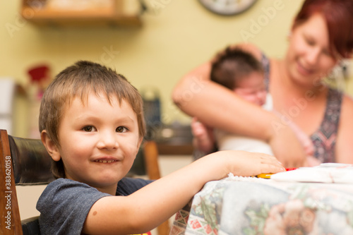 Young child sitting at table with mother and baby in background