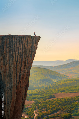 A small figure of a woman tourist with open arms, standing on the edge of a cliff in the summer mountains. The concept of a small man in the midst of a powerful nature.