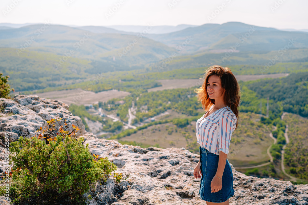 A beautiful woman stands on a high cliff, enjoying the view of the mountain range and forest