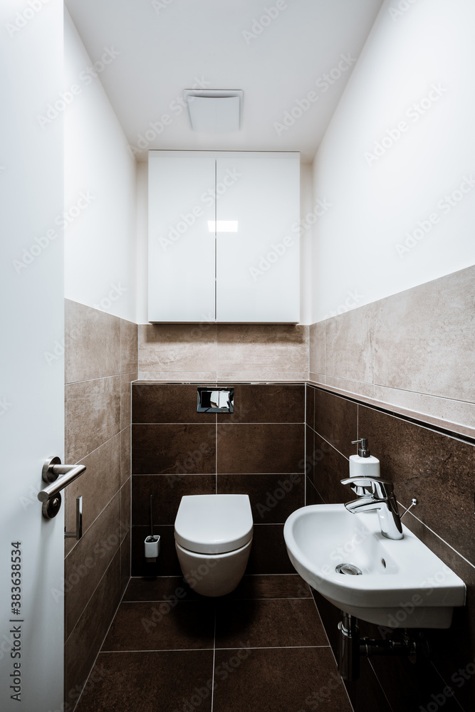 Small modern restroom with all furnishings, including a small sink, cabinet and ceiling fan. Paving and tiling have texture of concrete trowel in shade of brown. The toilet seat is closed. Small modern restroom with all furnishings, including a small sink, cabinet and ceiling fan. Paving and tiling have texture of concrete trowel in shade of brown. The toilet seat is closed.
