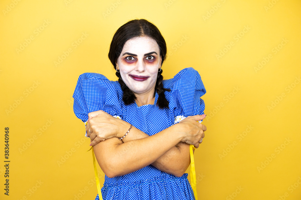 Obraz premium woman wearing a scary doll halloween costume over yellow background hugging oneself happy and positive, smiling confident