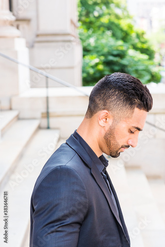 Portrait of Young East Indian American Businessman with beard in New York City, wearing black suit, black shirt, standing on stairs outside office building, looking down, thinking, lost in thought..