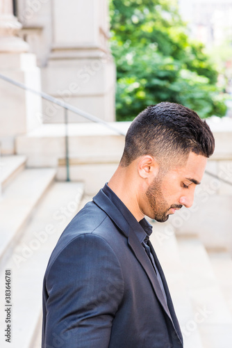 Portrait of Young East Indian American Businessman with beard in New York City, wearing black suit, black shirt, standing on stairs outside office building, looking down, thinking, lost in thought.. © Alexander Image