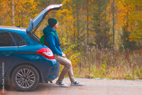 A man sits on the trunk of his car in the woods. Male camping in the forest, autumn landscape. Copy Space