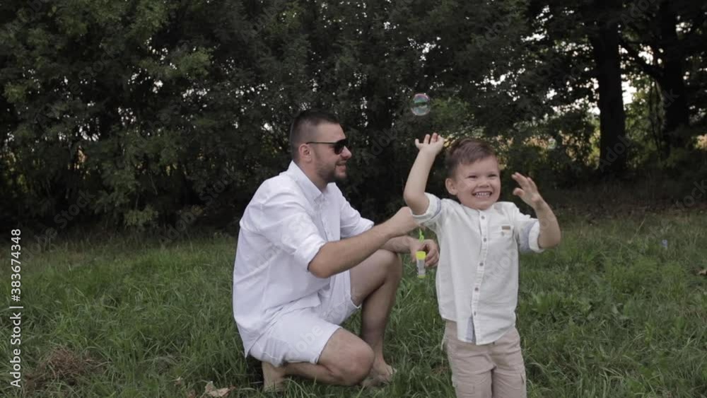 A handsome man with short dark hair in a white shirt, shorts crouched on one knee and plays with a little boy in the park in the summer A handsome man with short dark hair in a white shirt, shorts crouched on one knee and plays with a little boy in the park in the summer
