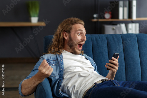 Curly-haired guy in casual style, watching great news on his mobile phone. Student receiving sms message reading good news. Surprised Man celebrating Victory on phone in Apartment.