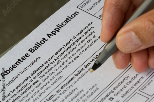 Hand holding a pen and filling out a New York State Absentee Ballot Application form. Selective focus on the form.