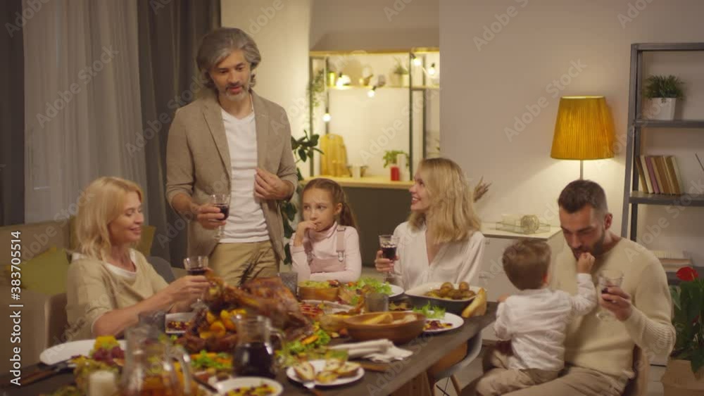 Mature man standing at table proposing toast then clinking glasses with family while celebrating Thanksgiving day Mature man standing at table proposing toast then clinking glasses with family while celebrating Thanksgiving day