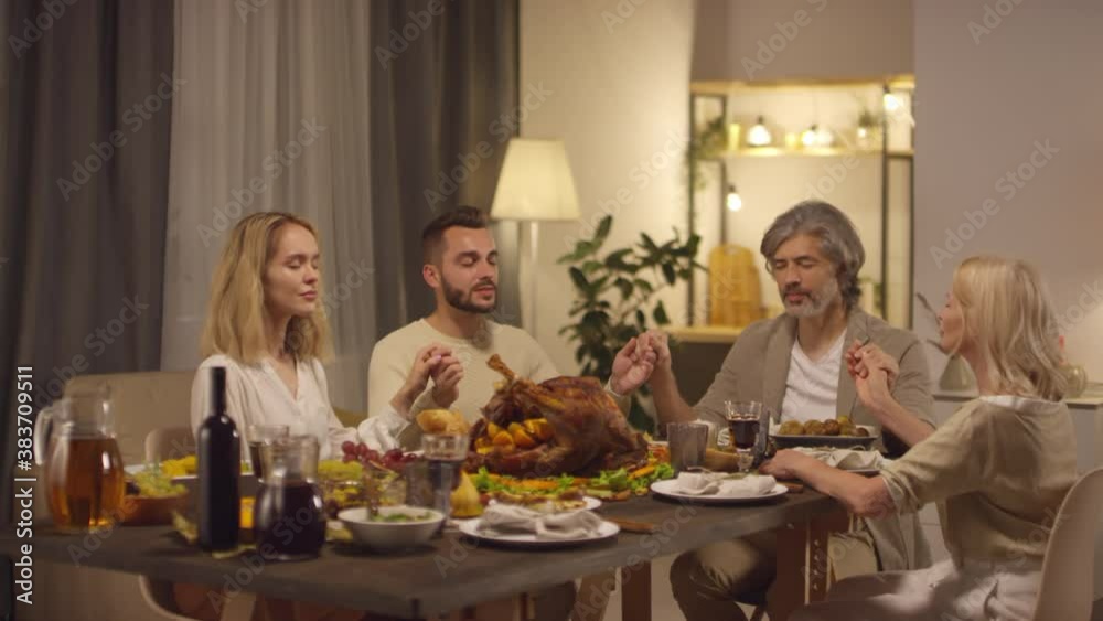 Family of four sitting at beautiful dining table with eyes closed, holding hands and praying to God on Thanksgiving day Family of four sitting at beautiful dining table with eyes closed, holding hands and praying to God on Thanksgiving day