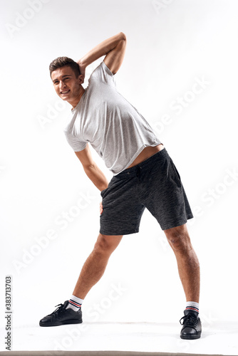 A sports man in shorts and a full-length T-shirt does exercises on a light background
