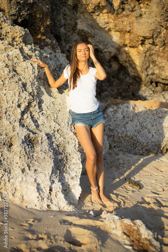 Slim young woman standing barefoot on the sand near the rock. Caucasian woman wearing jeans shorts, white T-shirt. Enjoying sunlight at the beach. Vacation in Asia. Travel lifestyle. Bali