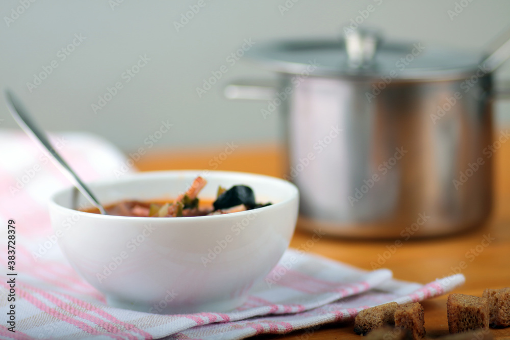 white bowl of thick soup of vegetables and meat, croutons and saucepan on white napkin on white wooden background white bowl of thick soup of vegetables and meat, croutons and saucepan on white napkin on white wooden background