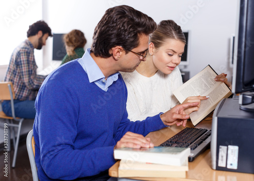 Young people preparing for exam in college library, reading book and using pc for searching information