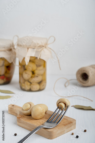 Marinated or fermented champignon mushrooms served on cutting board with fork against glass can, black pepper and bay leaf on white wooden background at kitchen. Vertical orientation image
