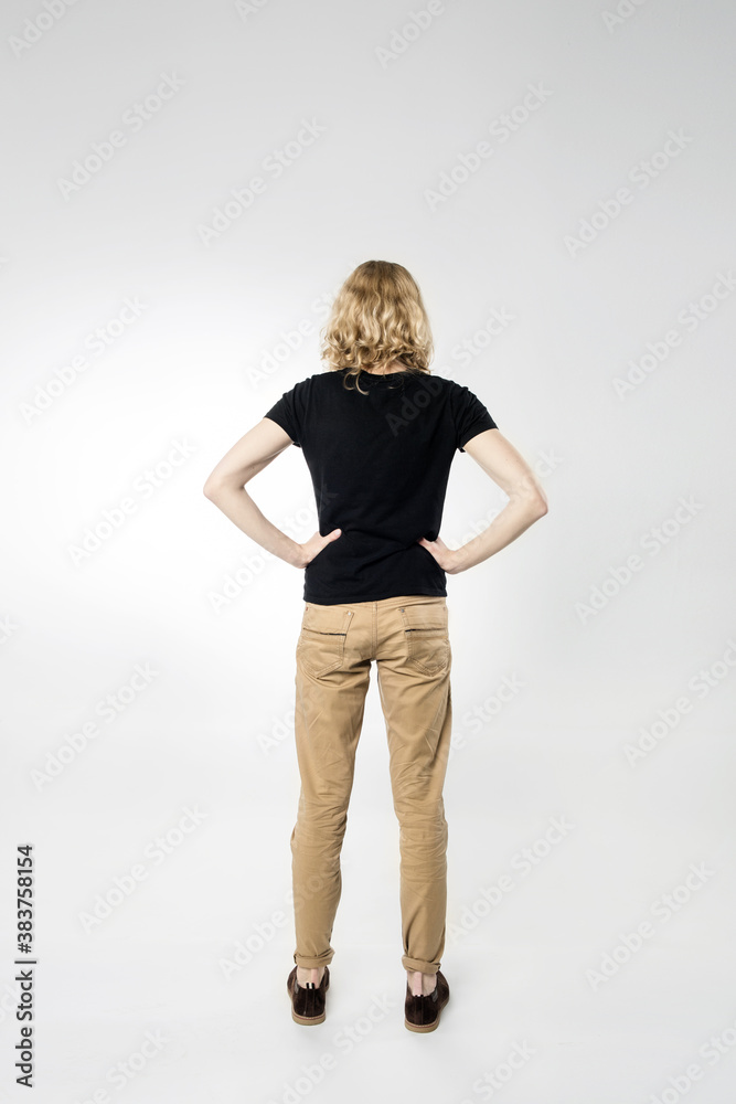 A thin young man with long blonde hair in a black T shirt and beige jeans, stands with back to the camera with his hands on his waist. Isolated on a light background. A thin young man with long blonde hair in a black T shirt and beige jeans, stands with back to the camera with his hands on his waist. Isolated on a light background.