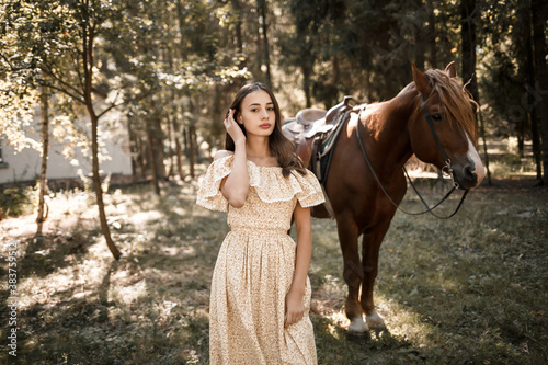 A beautiful young girl dressed in a dress stands near a horse in the forest