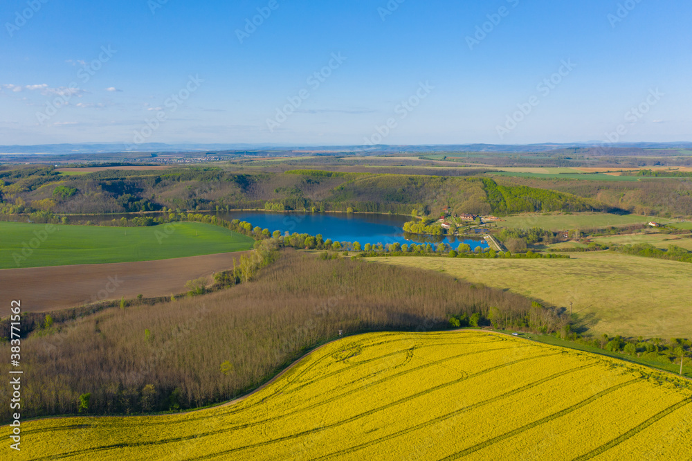 Notincs, Hungary - Aerial view of cultivated blooming canola field at countryside with tiny lake at the background and beautiful spring colors. 