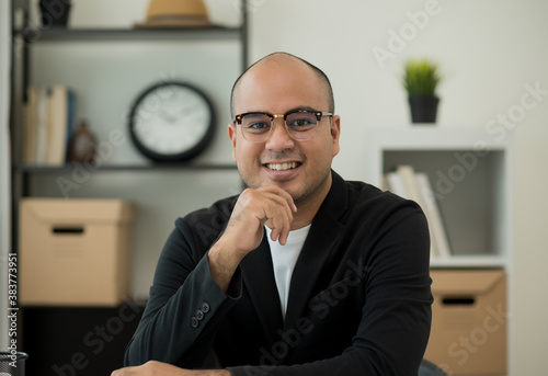 A middle-aged man around the age of 35. Working at home looking at camera. He was wearing a black suit and glasses. Smiling asian businessman work from home.