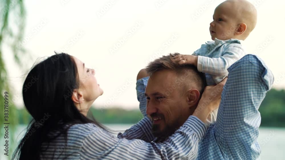Woman tickling baby sitting on shoulders of man. Happy parents playing with little child near river. Concept of family Woman tickling baby sitting on shoulders of man. Happy parents playing with little child near river. Concept of family
