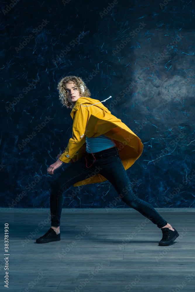 A young guy with a surfer's hair in a yellow jacket and headphones is dancing against a dark background