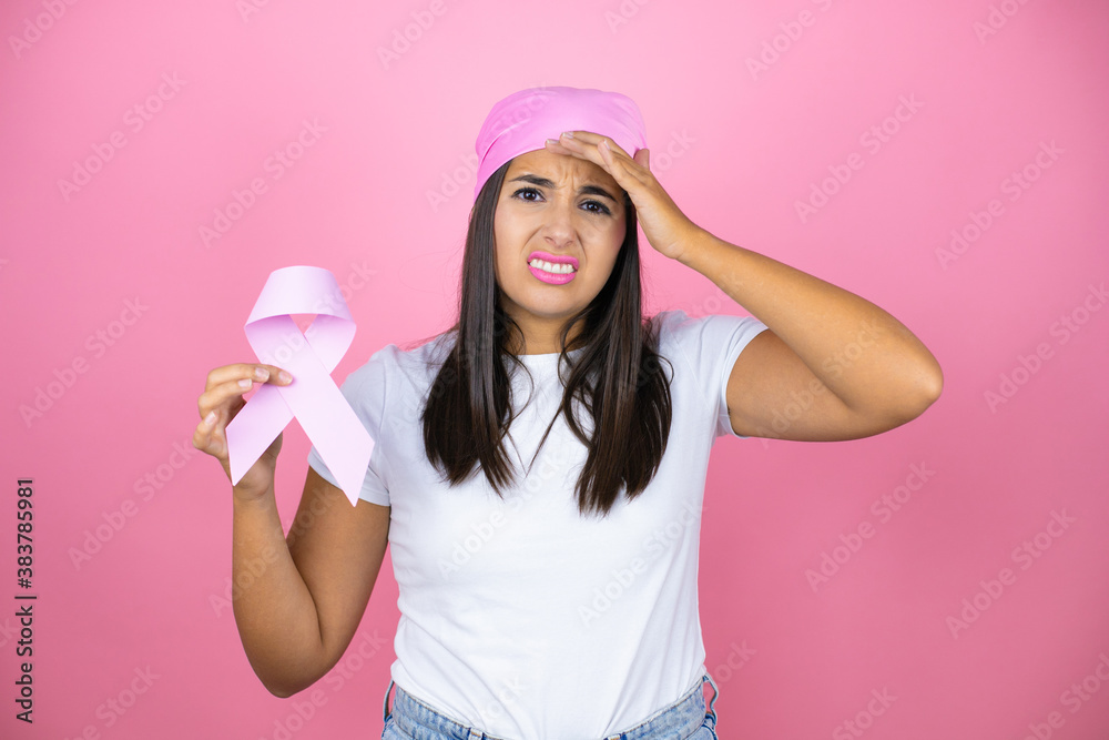 Young beautiful woman wearing pink headscarf holding brest cancer ribbon over isolated pink background stressed and frustrated with hand on head, surprised and angry face Young beautiful woman wearing pink headscarf holding brest cancer ribbon over isolated pink background stressed and frustrated with hand on head, surprised and angry face