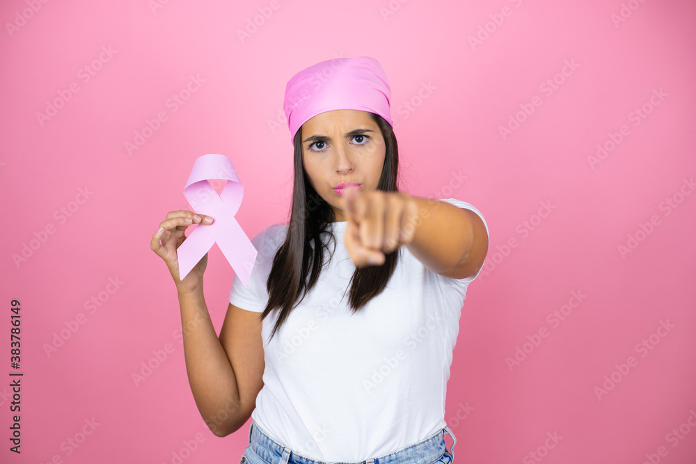 Young beautiful woman wearing pink headscarf holding brest cancer ribbon over isolated pink background pointing with finger to the camera and to you, hand sign, gesture from the front Young beautiful woman wearing pink headscarf holding brest cancer ribbon over isolated pink background pointing with finger to the camera and to you, hand sign, gesture from the front