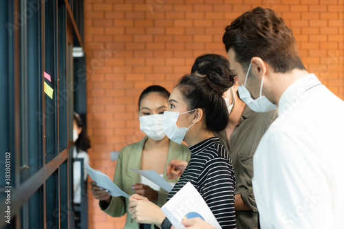 Young Asian businesswoman in protective face mask looking at a sticky note on office glass window after she and her colleagues brainstormed on new business plan and write down the strategic plan words