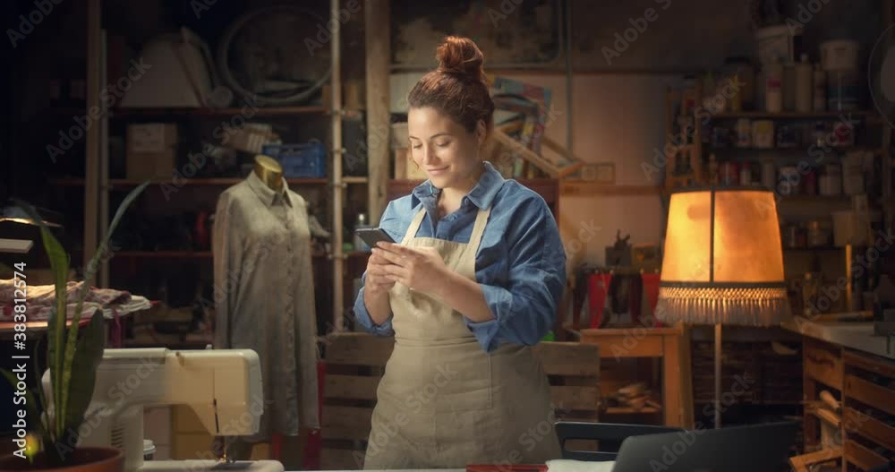 Attractive young ginger woman chatting online connected in app with smartphone indoors, beautiful happy female smiling standing next to work-desk using mobile device at creative design studio