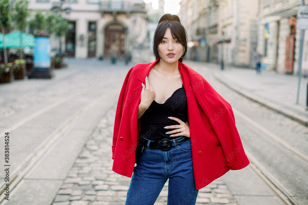Elegant pretty Asian lady with black long ponytail hair, wearing stylish red jacket, black top and jeans, posing to camera at summer day morning in the old city street. Urban street portrait