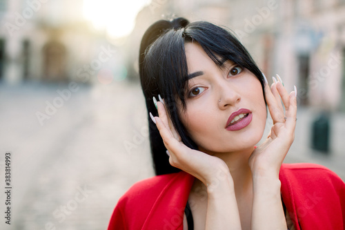 Lovely young Asian woman, wearing red blazer, with black long ponytail hairstyle, posing on the street of old European city on sunny day. Close-up face portrait, copy space