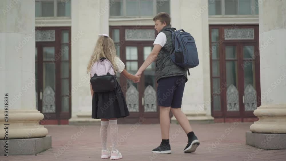 Wide shot back view of Caucasian brother leading sister to school. Little girl not willing to study as boy pulling her hand. Siblings walking to study in the morning. Education concept. Wide shot back view of Caucasian brother leading sister to school. Little girl not willing to study as boy pulling her hand. Siblings walking to study in the morning. Education concept.