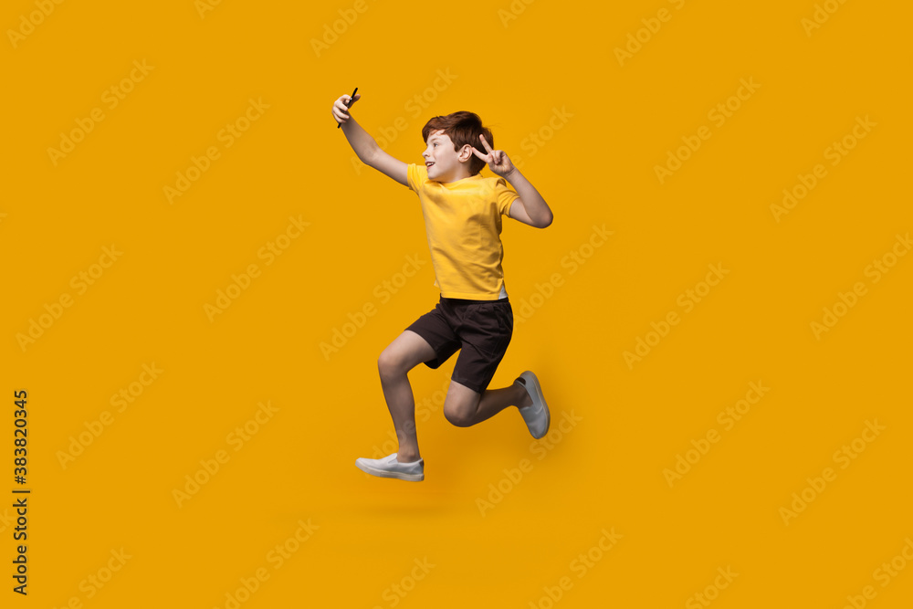 Ginger caucasian boy jumping on a yellow studio wall and making a selfie using a phone gesturing the piece sign and smile