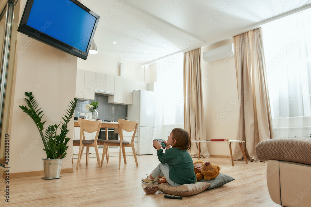 A little cute girl holding a gamepad playing videogames on a TV while sitting on a floor cross-legged A little cute girl holding a gamepad playing videogames on a TV while sitting on a floor cross-legged