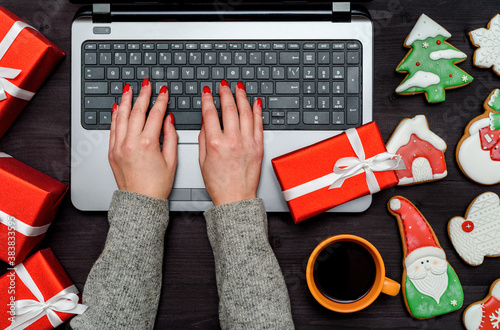 Top view of woman hands typing on open laptop keyboard on table with Cristmas decorations (gift boxes and gingerbread cookies), copy space. Flat lay photo