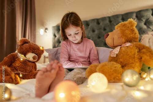 A cute girl wearing pyjamas sitting alone on a huge bed barefoot watching cartoons on a tablet before going to sleep with her fluffy toys and garlands all over the bed