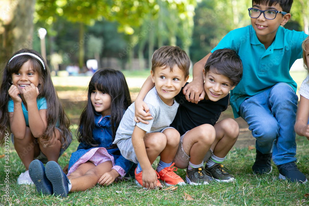 Cheerful kids sitting and squatting down on grass, hugging each other, looking away in excitement. Kids play or entertainment concept Cheerful kids sitting and squatting down on grass, hugging each other, looking away in excitement. Kids play or entertainment concept