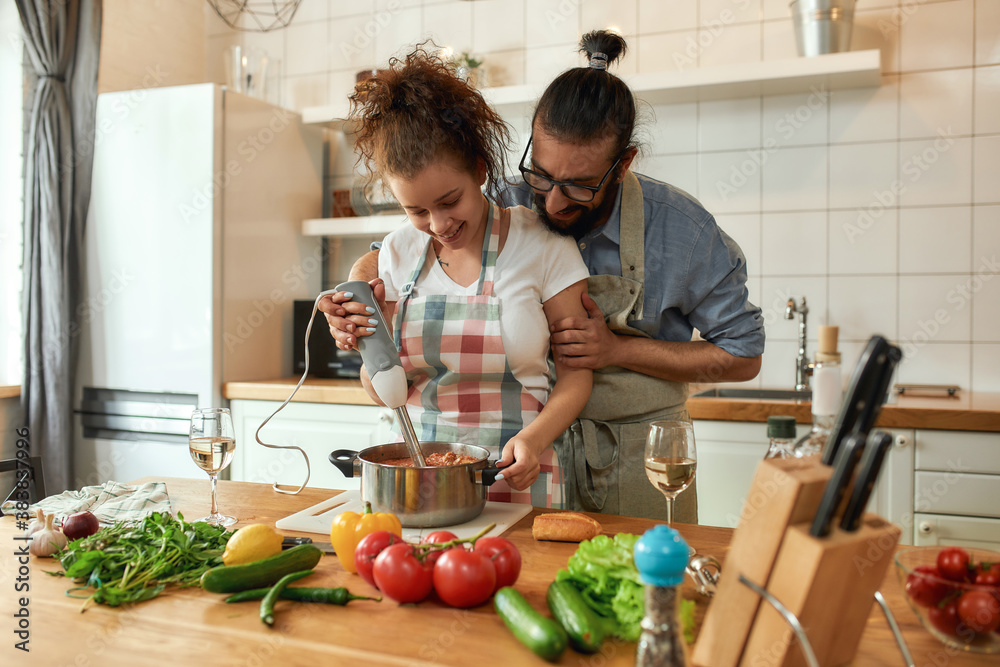 © Svitlana - Young couple preparing a meal together in the kitchen. Italian man, chef cook helping his girlfriend to use hand blender. Cooking at home, Italian cuisine