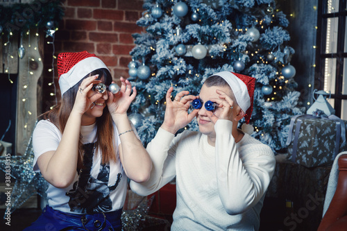 A young man and woman in Santa Claus hats are fooling around on the background of the New Year tree, closed their eyes with Christmas toys in the shape of a ball and smiling.