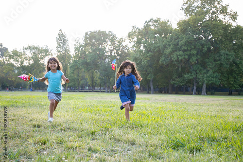 Joyful black haired little girls with pinwheels running on grass  having race in park. Front view  full length. Children outdoor activity concept