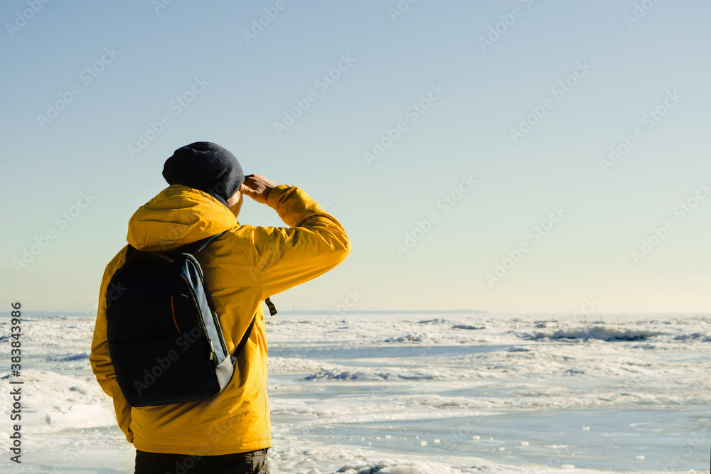 Obraz premium One faceless man in yellow raincoat with backpack standing on frozen sea and looking far on nature. Copy space. Tourist hiking. Freedom of travel concept. Unrecognizable male