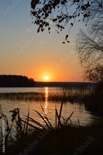 Sunset on the lake. Orange glow and darkening shore with trees and plants.